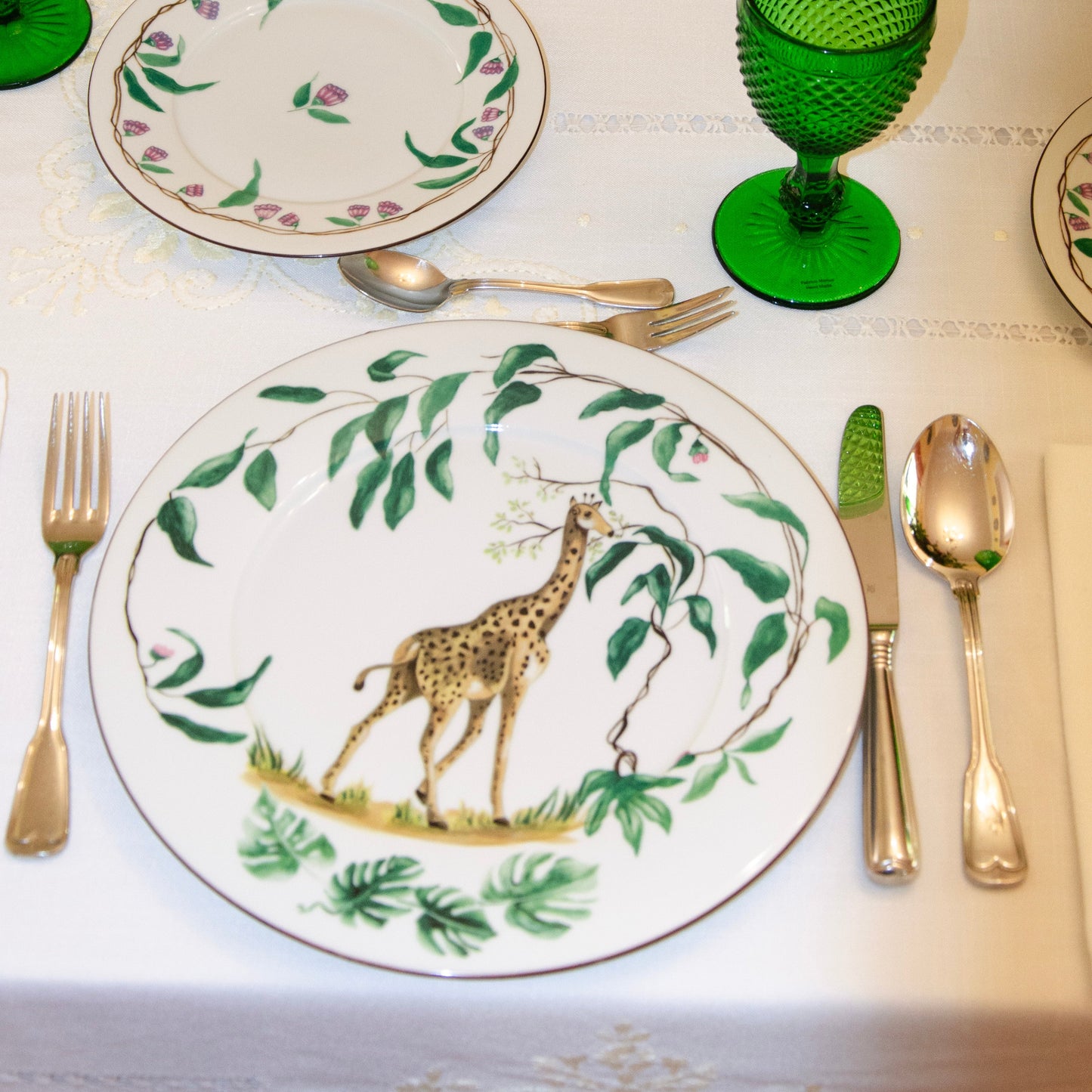 Dinner table setting with decorative plates featuring giraffes, green glasses, and cutlery.